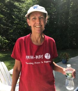 Diane standing outside holding a water bottle wearing a path-way hat and red ReImagned Picnic T-shirt smiling at the camera.