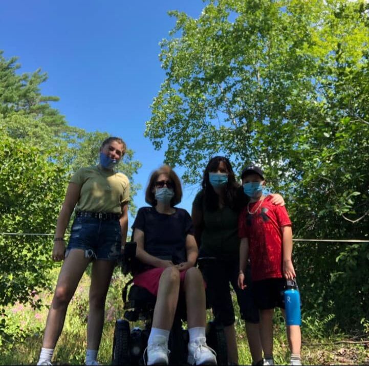 Donna sitting in her power chair with 3 friends standing next to her outside.All are looking at the camera wearing masks.