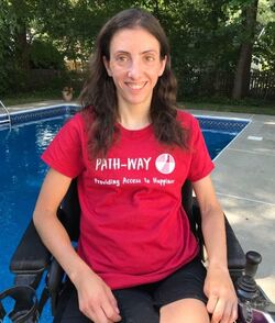 Steph sitting in her power chair outside in front of a pool wearing a  red ReImagned Picnic T-shirt smiling at the camera.