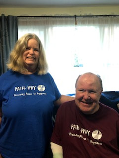 Susan wearing a blue ReImagined Picnic T standing next to Bob wearing a maroon ReImagined picnic t sitting in his power chair. Both are smiling at the camera.